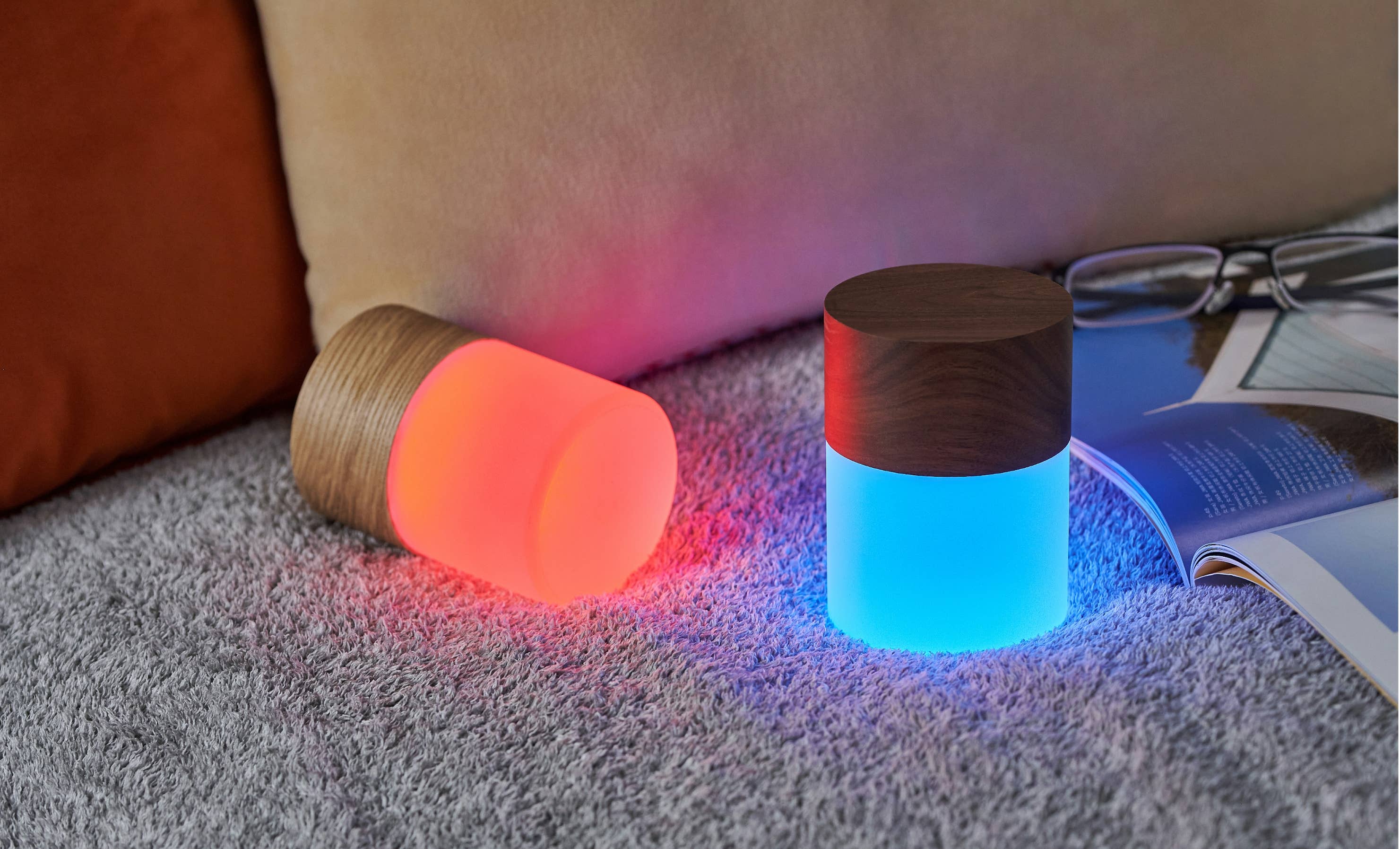 Two Lemelia lights on grey carpet beside a book, glowing coral and blue