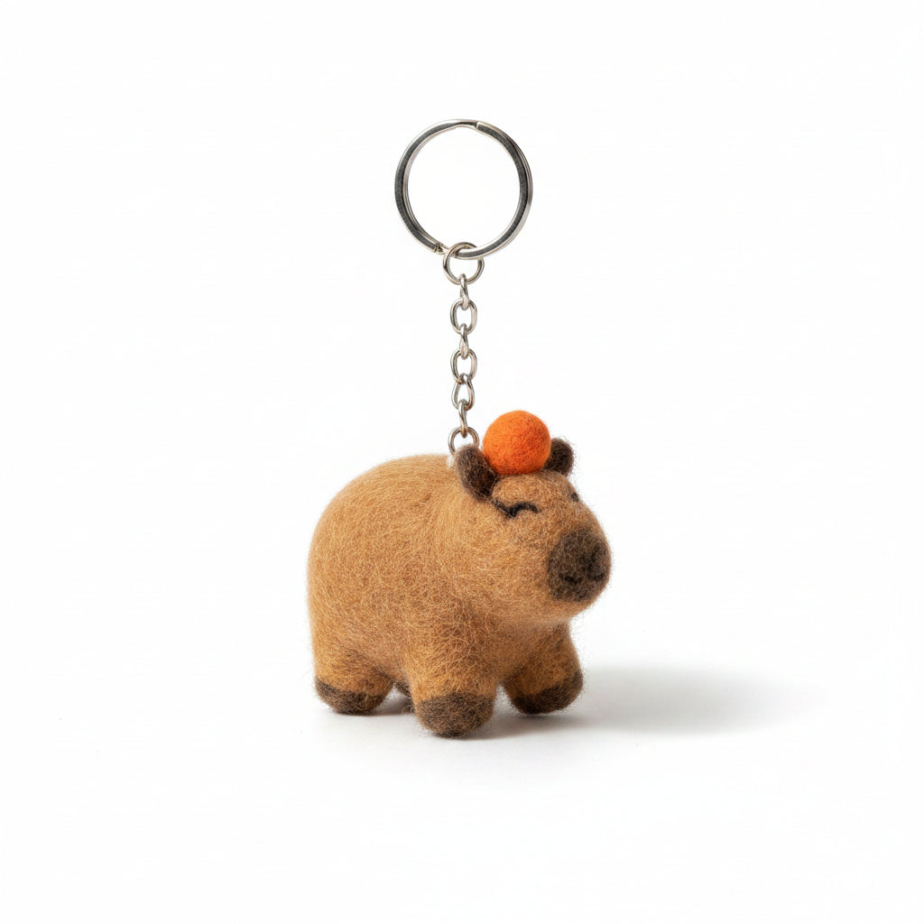 A small felted animal keychain with a warm brown body, dark nose and feet, and an orange accent on top, hanging from metal keys held by a person's hand on a wooden table.