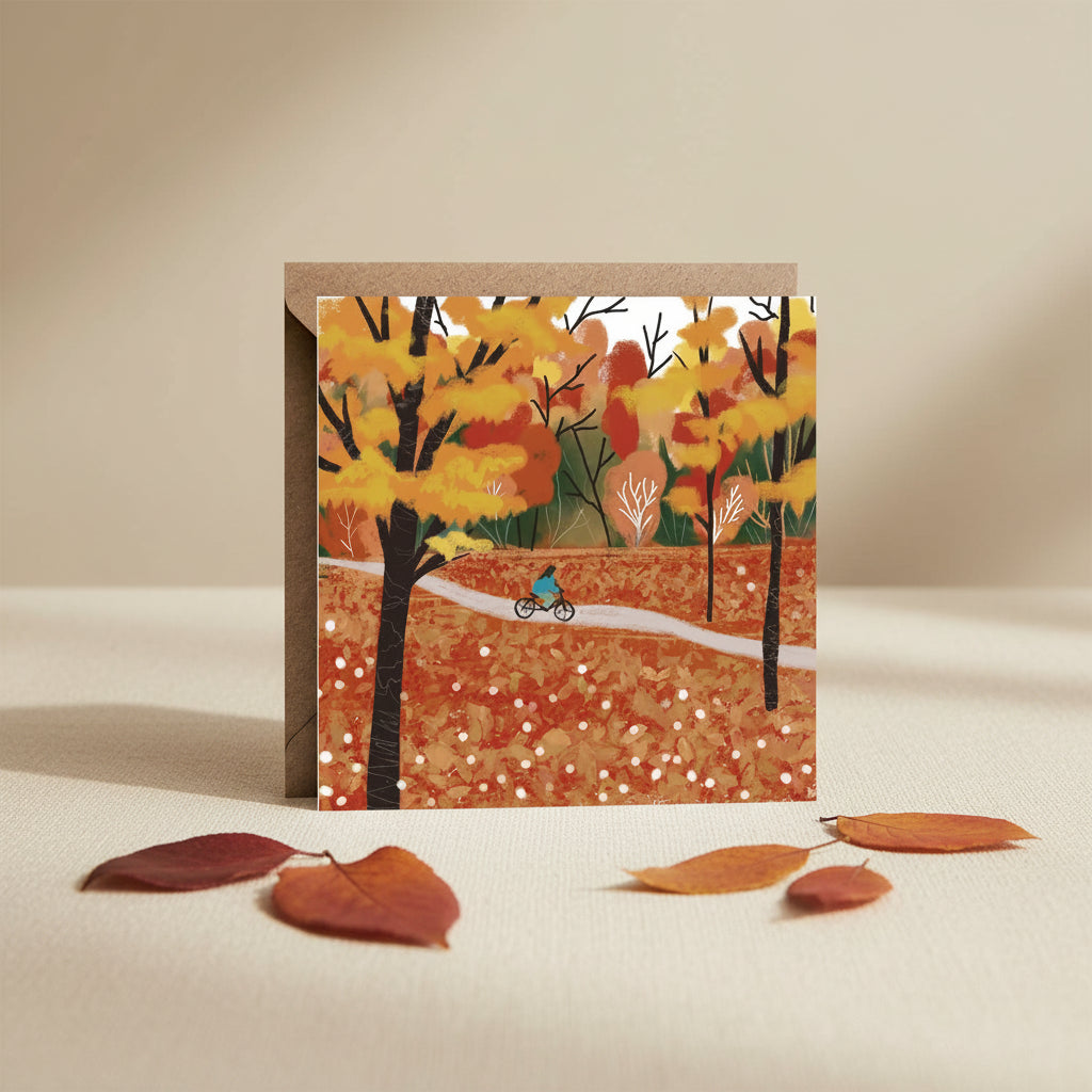 An autumn-themed art print or card featuring a cyclist on a path through a forest of orange, yellow, and red trees, shown with a brown kraft envelope behind it.