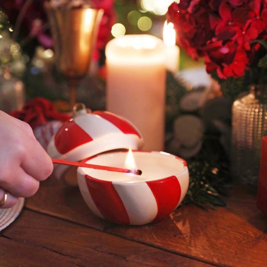 Hand lighting Candy Cane Stripe Cinnamon Bauble Candle on a festive dinner table with candles and red flowers.