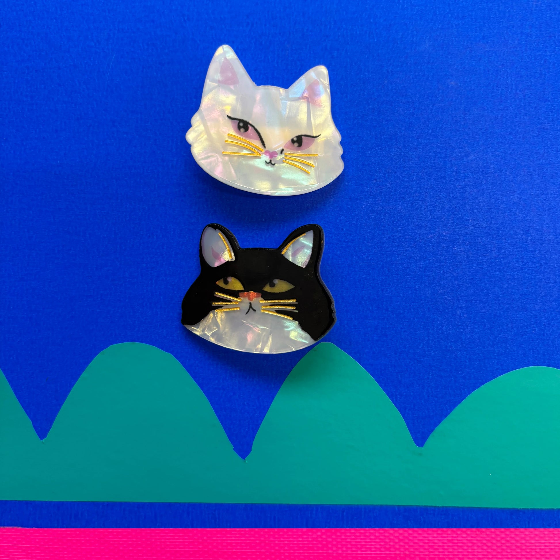 Flat lay of two cat-shaped hair clips—one white and one black—against a vibrant blue background with green and pink geometric accents.