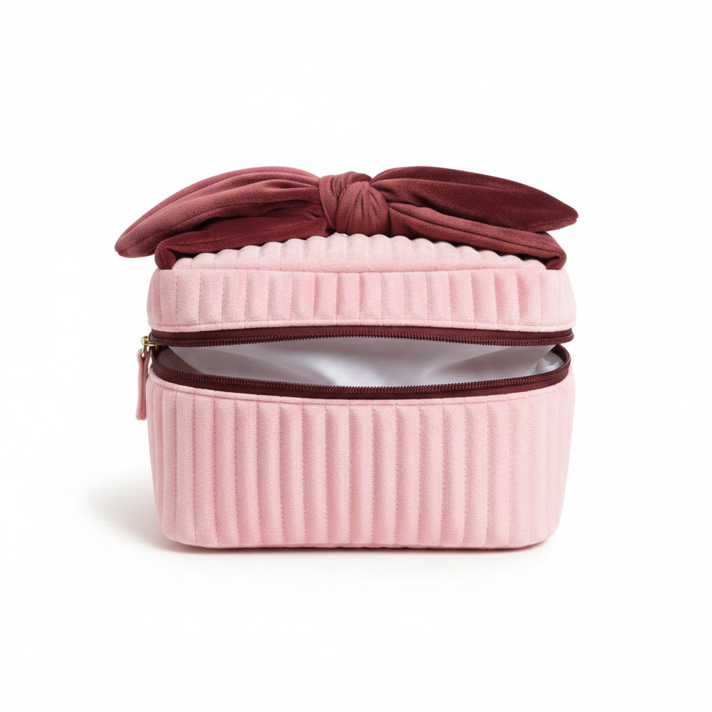 Pale pink velvet makeup bag with burgundy bow handle and gold zip on cream background.