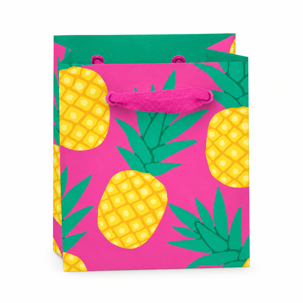 Mini pineapple gift bag with bright yellow fruit and green leaves on a bold pink background, featuring matching pink grosgrain ribbon handles and a gift tag. Made from recyclable FSC-certified paper.
