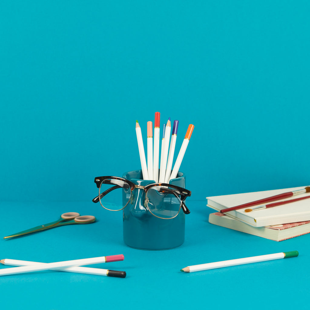 Teal nose pen holder with pencils, scissors and notebooks on a turquoise desk