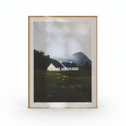 Framed painting of a white cottage in a landscape with mountains on a beige wall.