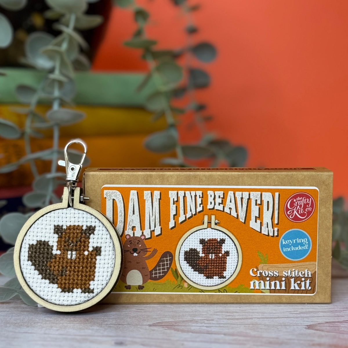 Beaver cross stitch keyring beside its matchbox in front of an orange wall