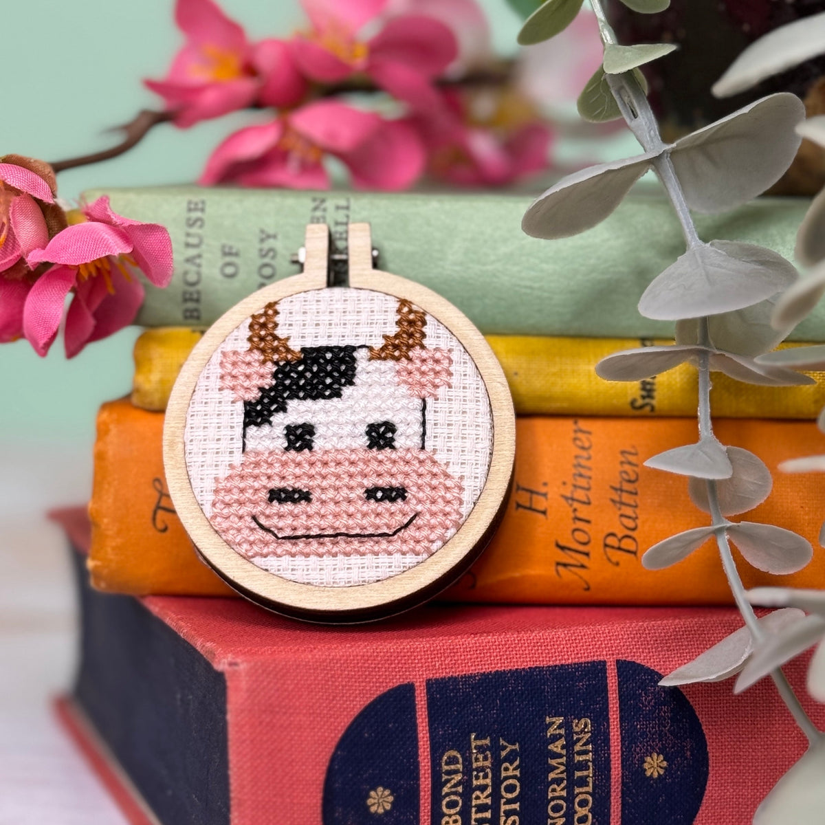 Cow face cross stitch hoop propped on a stack of books with pink flowers
