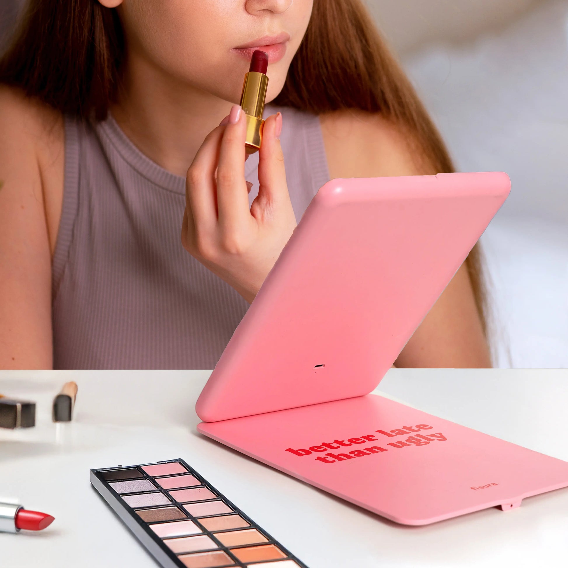 Person applying lipstick using the pink LED mirror beside a makeup palette