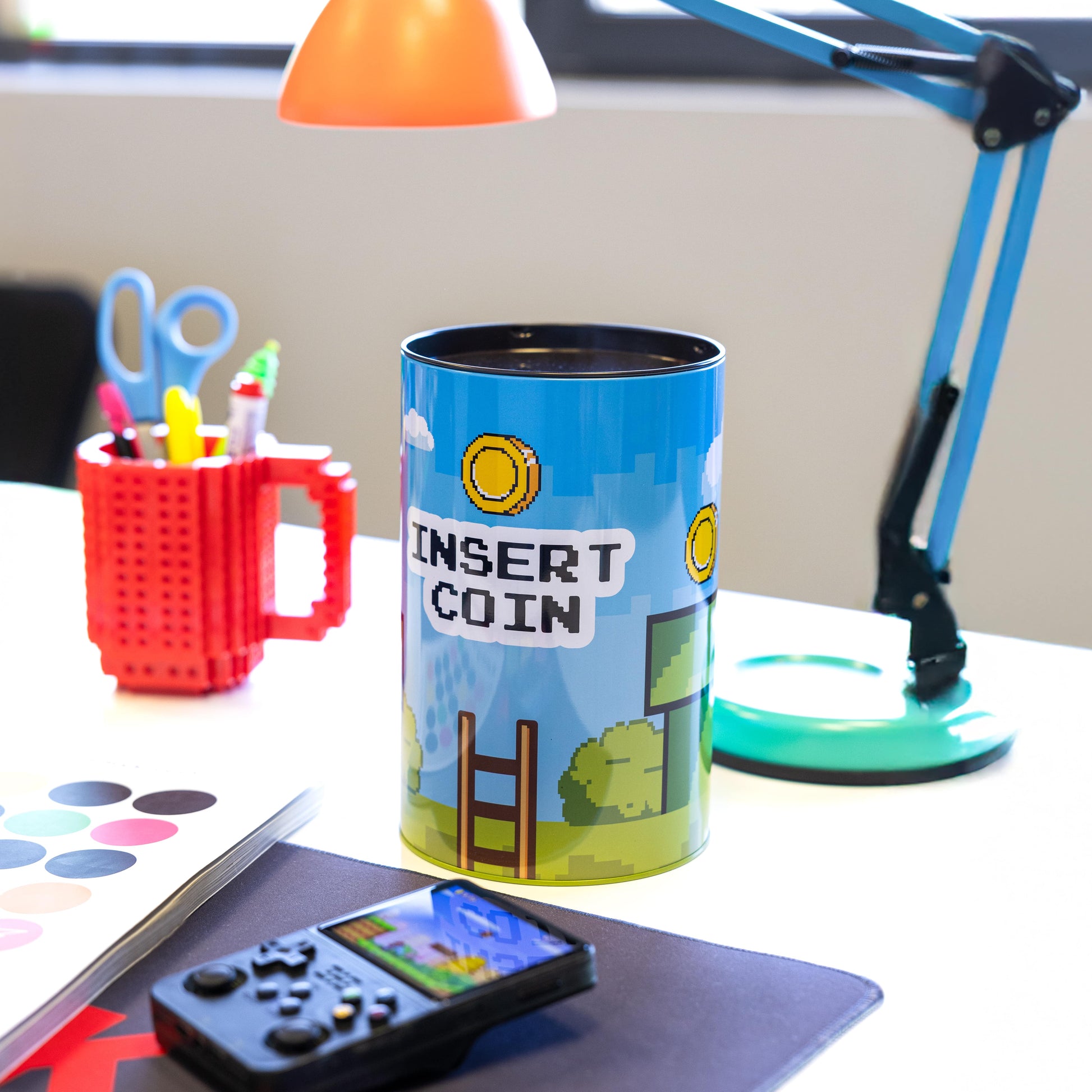Insert Coin money tin on a desk beside a games controller and mug