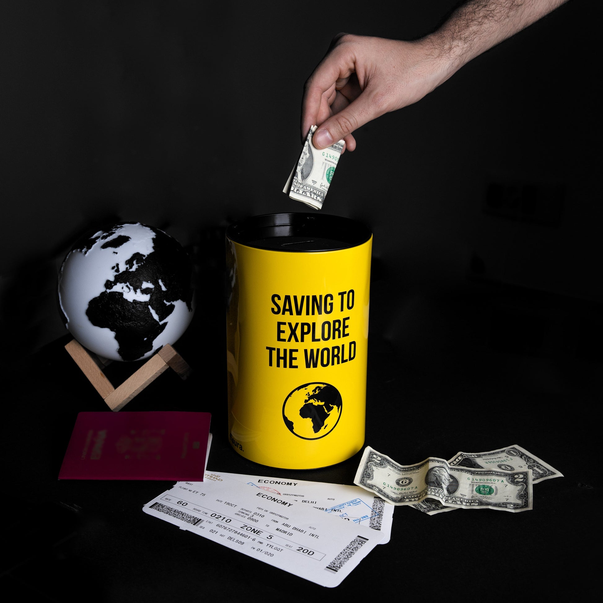 Hand posting a banknote into the yellow travel tin beside a globe