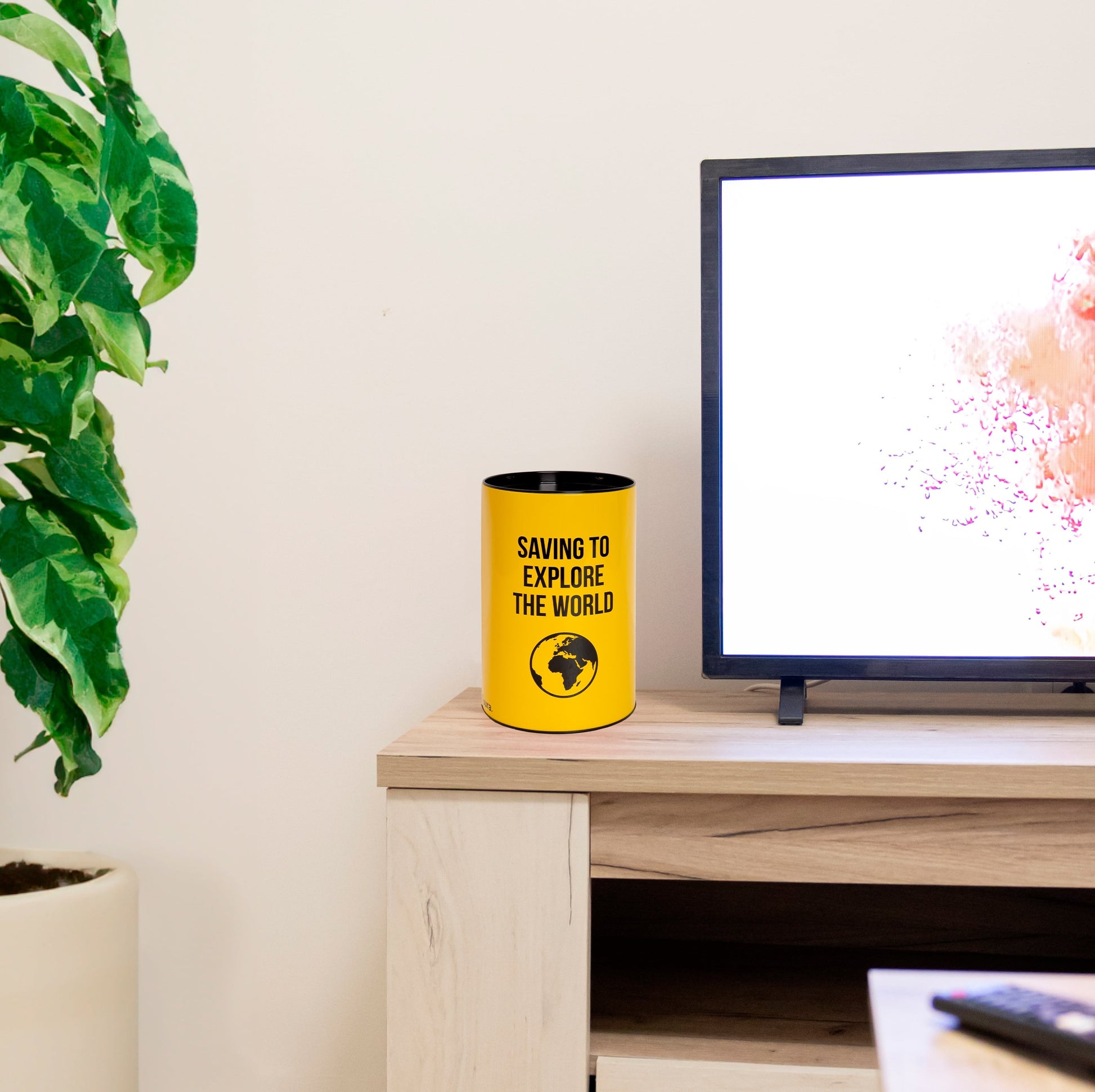 Yellow savings tin on a light wooden desk beside a monitor and plant