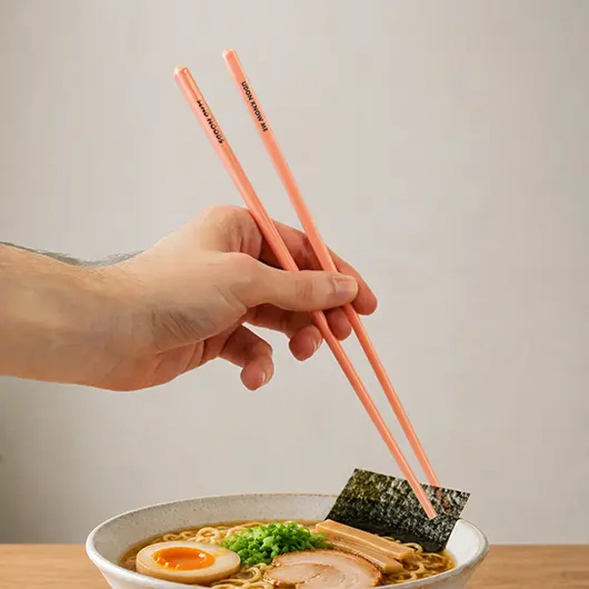 Hand using pink ceramic chopsticks to lift nori over a ramen bowl