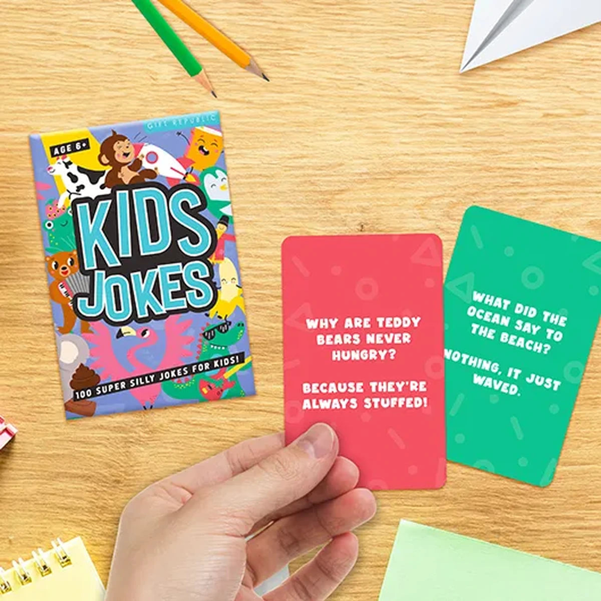 Hand holding the kids jokes pack beside scattered joke cards