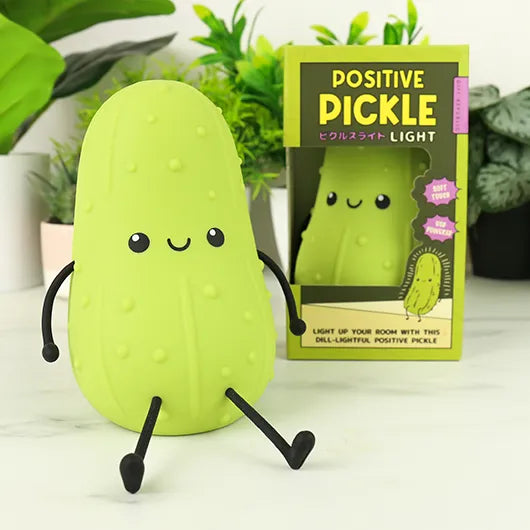 Smiling pickle lamp sitting beside its packaging with plants behind