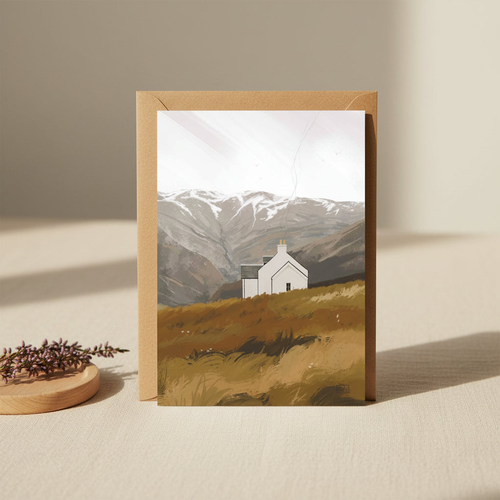A greeting card with a kraft paper envelope, featuring an illustration of a small white cottage on a grassy hill with mountains in the background.