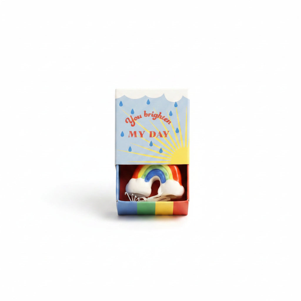Ceramic rainbow inside open blue matchbox with sunshine and raindrop design on cream background