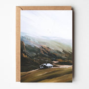Mountain Calm greeting card featuring an illustrated cottage in a soft Norwegian mountain landscape with misty hills and earthy tones, paired with kraft envelope.