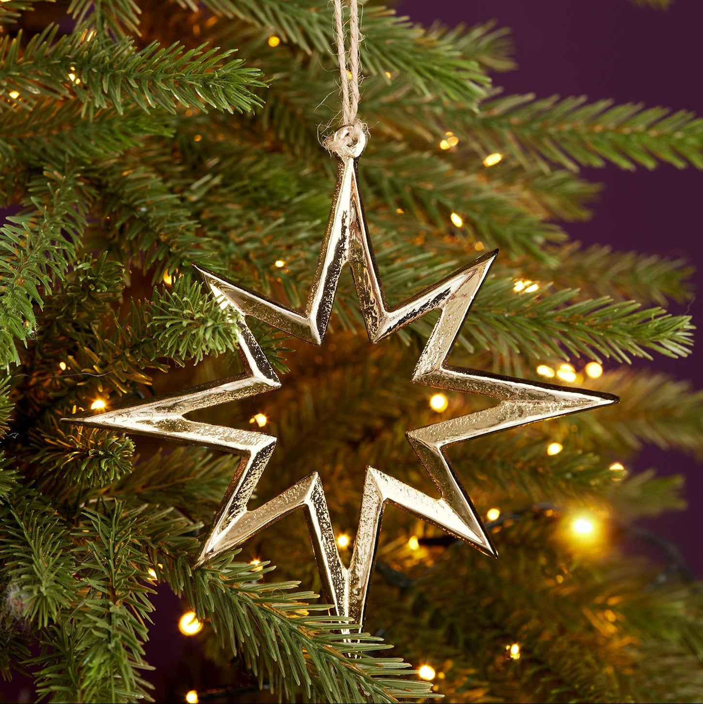 A metallic silver star-shaped Christmas ornament hanging from a green Christmas tree branch with warm string lights in the background.