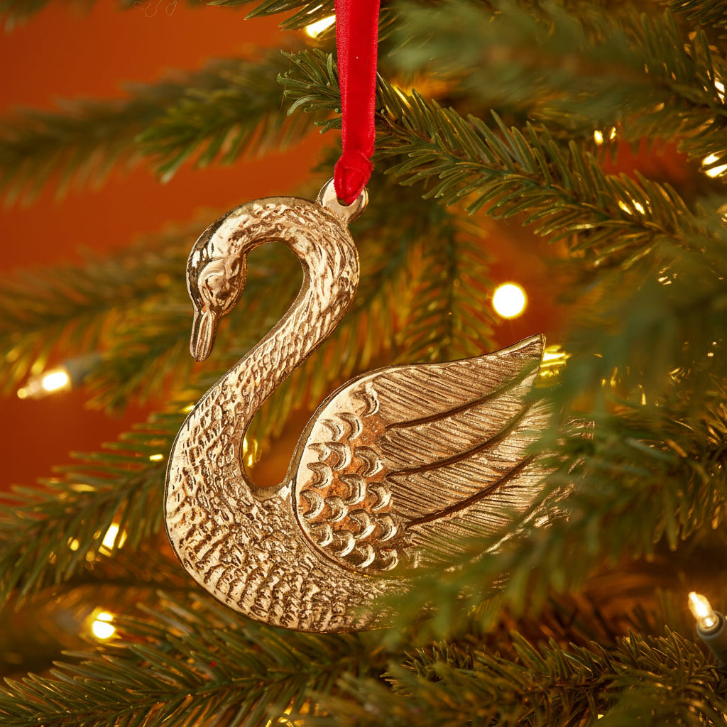 A metallic gold-toned swan Christmas ornament hanging from a red ribbon on a decorated Christmas tree with warm lights.