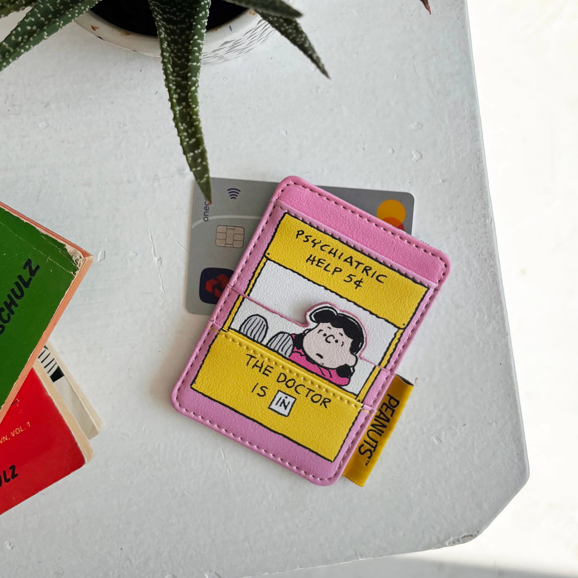 Peanuts Lucy Cardholder with “Psychiatric Help 5¢” yellow booth design and Lucy illustration on white table beside Peanuts books.