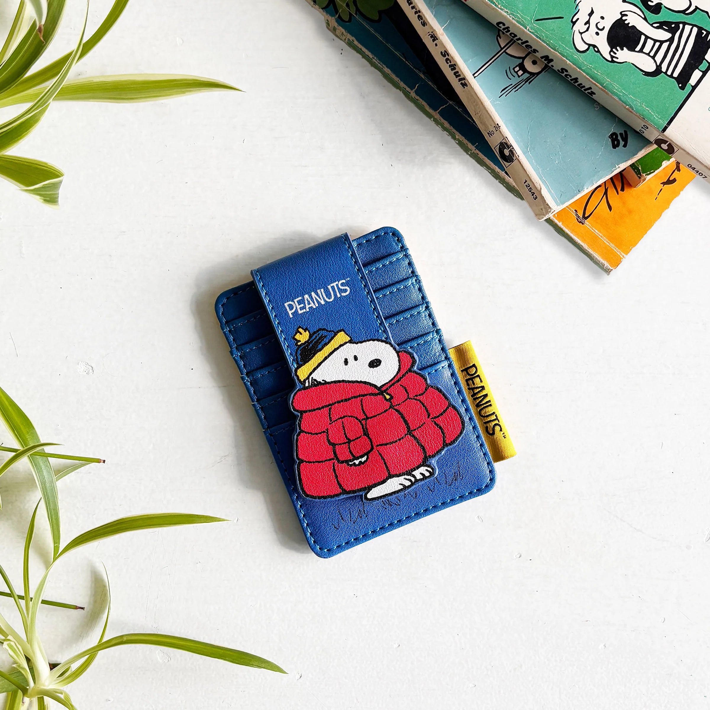 Peanuts Snoopy Coat Foldover Card Holder in blue with Snoopy wearing a red puffer coat and yellow hat, styled beside Peanuts comic books.