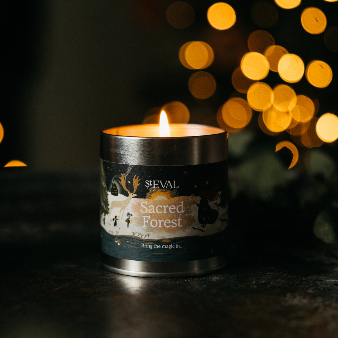 Sacred Forest Christmas Tin Candle lit on a dark table with festive lights behind