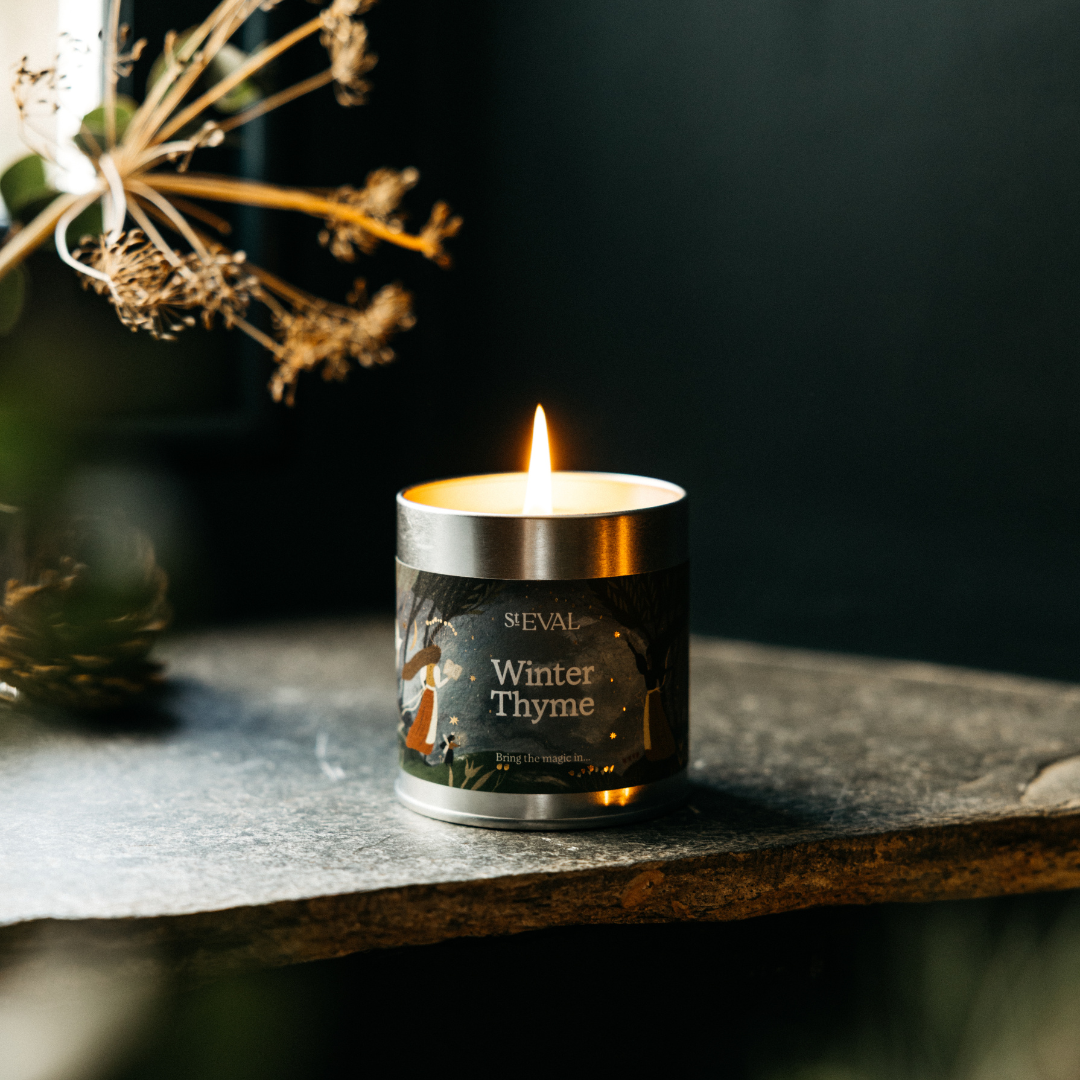 Winter Thyme Christmas Tin Candle lit on a dark table with festive lights behind