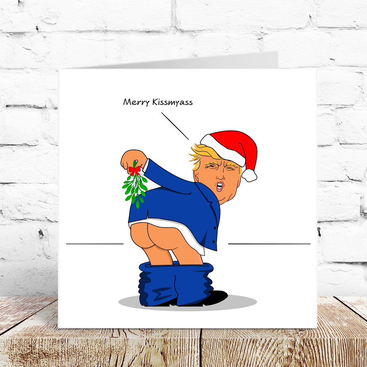 Trump Merry Kissmy*ss Christmas Card propped against a white brick wall