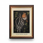 Framed art print featuring a skeleton hand holding a robin bird against a dark background with ivy leaves. The print has a cream mat and the text 'Kitty Willow Wilson' at the bottom, set in a dark wood frame.