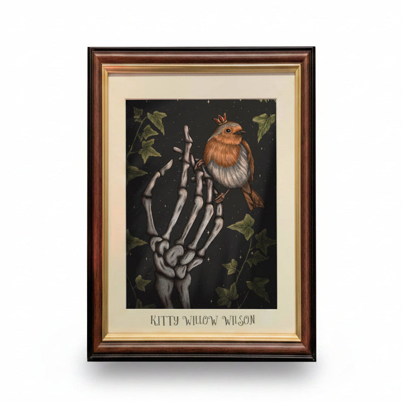 Framed art print featuring a skeleton hand holding a robin bird against a dark background with ivy leaves. The print has a cream mat and the text 'Kitty Willow Wilson' at the bottom, set in a dark wood frame.